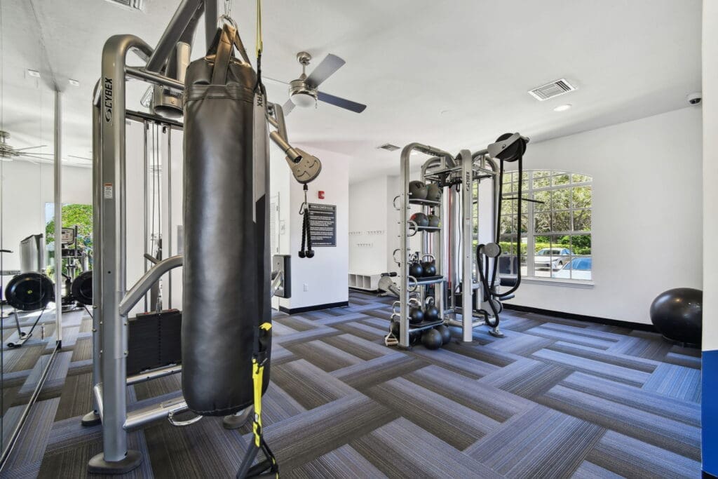 **Alt Text Description**: A well-equipped fitness center featuring a modern Cybex functional trainer with a heavy punching bag, cable machines, and a variety of kettlebells and medicine balls neatly stored on a rack. The flooring has a stylish dark gray and blue geometric pattern. Ceiling fans and large windows allow for ample natural light and ventilation. The space is clean, bright, and optimized for strength training and functional workouts. A sign on the wall outlines gym rules, and the area is designed to support a full-body workout experience.
