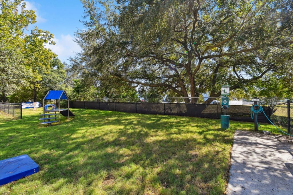 **Alt Text Description**: A fenced-in pet park shaded by large, mature trees, featuring a bright green grassy field. Within the park are several agility obstacles including a small platform with steps and a ramp, a blue tunnel, and a blue climbing structure with a roof. To the right, a pet waste station with a bag dispenser and trash bin is mounted on the fence next to a dog wash station equipped with a hose. A “Pet Park” sign is visible on the far fence, and the entire area is bathed in natural sunlight under a clear sky.