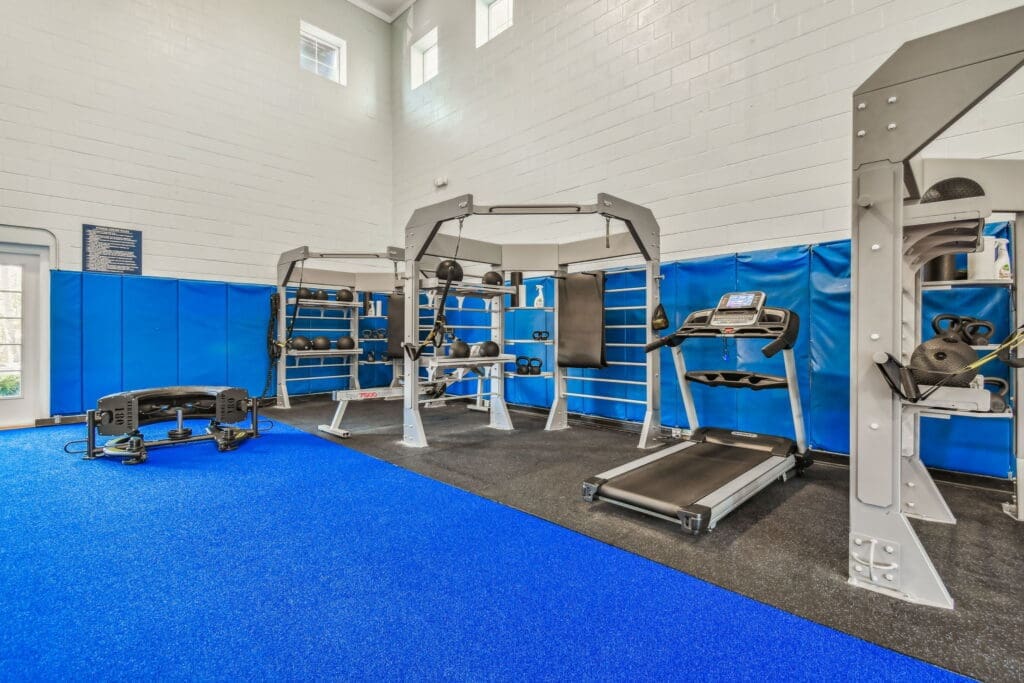 **Alt Text Description**: A well-equipped fitness center with high ceilings and natural light streaming through upper windows. The room features a variety of exercise equipment including a treadmill, a functional training rig, kettlebells, medicine balls, and a weight bench. The flooring is a combination of vibrant blue turf and black rubber, with blue padded wall panels surrounding the room for added safety. A doorway to the left leads outside, making the space bright and airy—ideal for strength training and functional workouts.