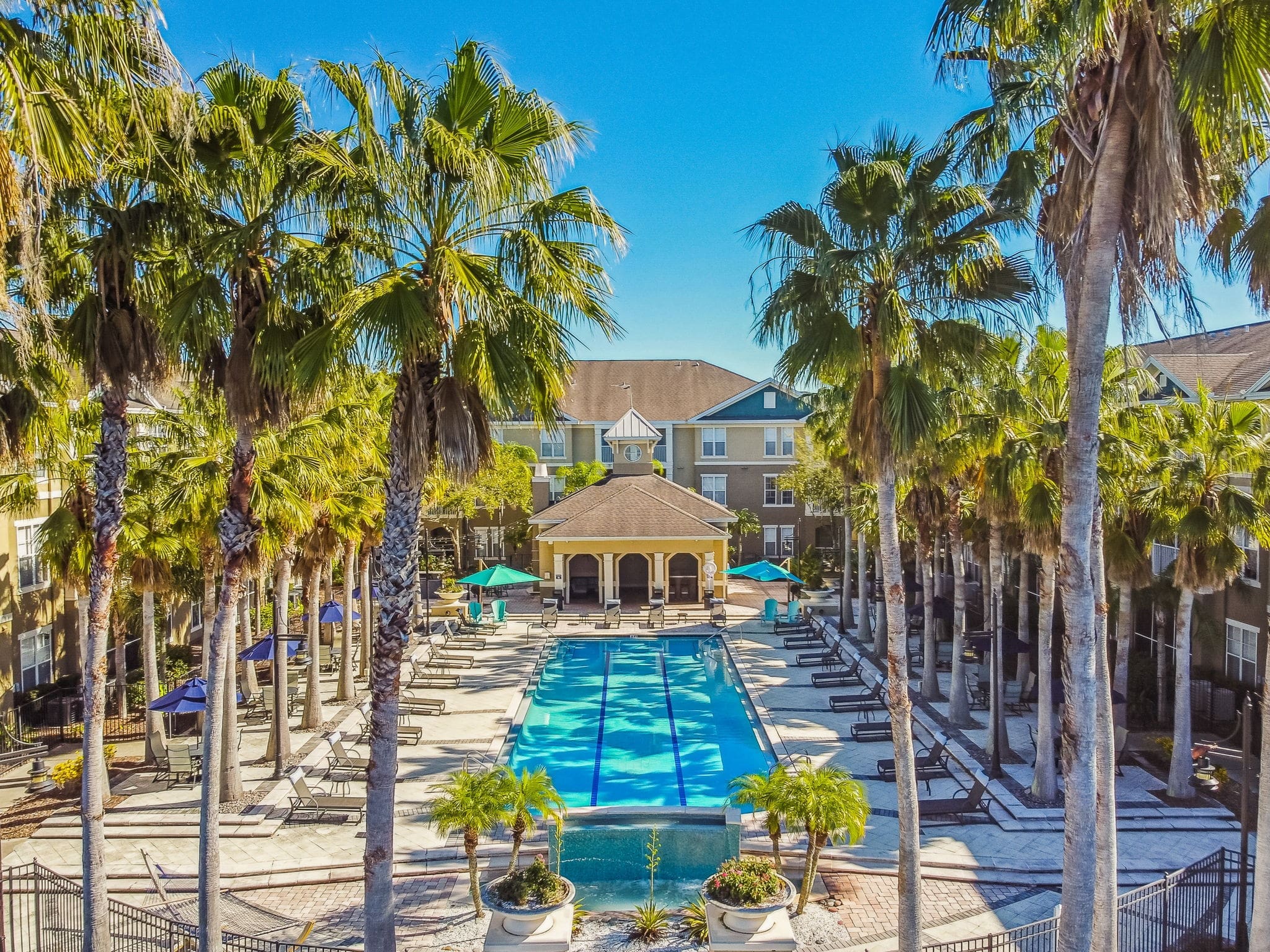 **Alt Text Description**: A luxurious outdoor swimming pool flanked by tall palm trees and surrounded by a three-story apartment complex. The rectangular pool is lined with neatly arranged lounge chairs and umbrellas in shades of blue and green on both sides. At the far end, a clubhouse with a peaked roof and arched entryway stands as a central focal point. The pool deck is paved with light-colored stone tiles, and decorative planters with palm trees and flowers enhance the resort-like ambiance. The sky is clear and blue, adding to the bright and inviting atmosphere.
