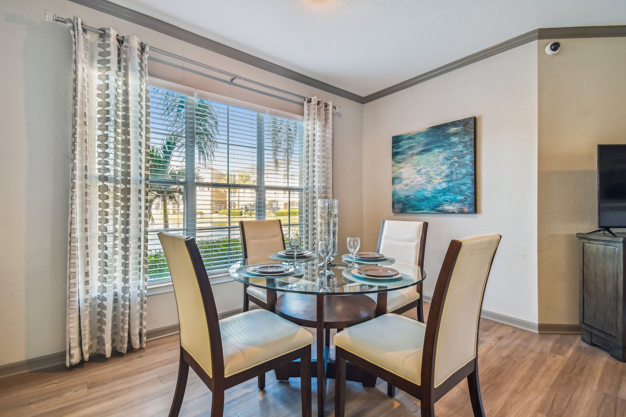 **Alt Text:** A bright and airy dining area in a modern apartment featuring a round glass-top table set for four with contemporary place settings. Four upholstered chairs with wood frames surround the table, which sits on wood-style flooring. Large windows with sheer, patterned curtains allow natural light to stream in, highlighting a teal and aqua abstract painting on the adjacent wall. A flat-screen TV is partially visible on the right.