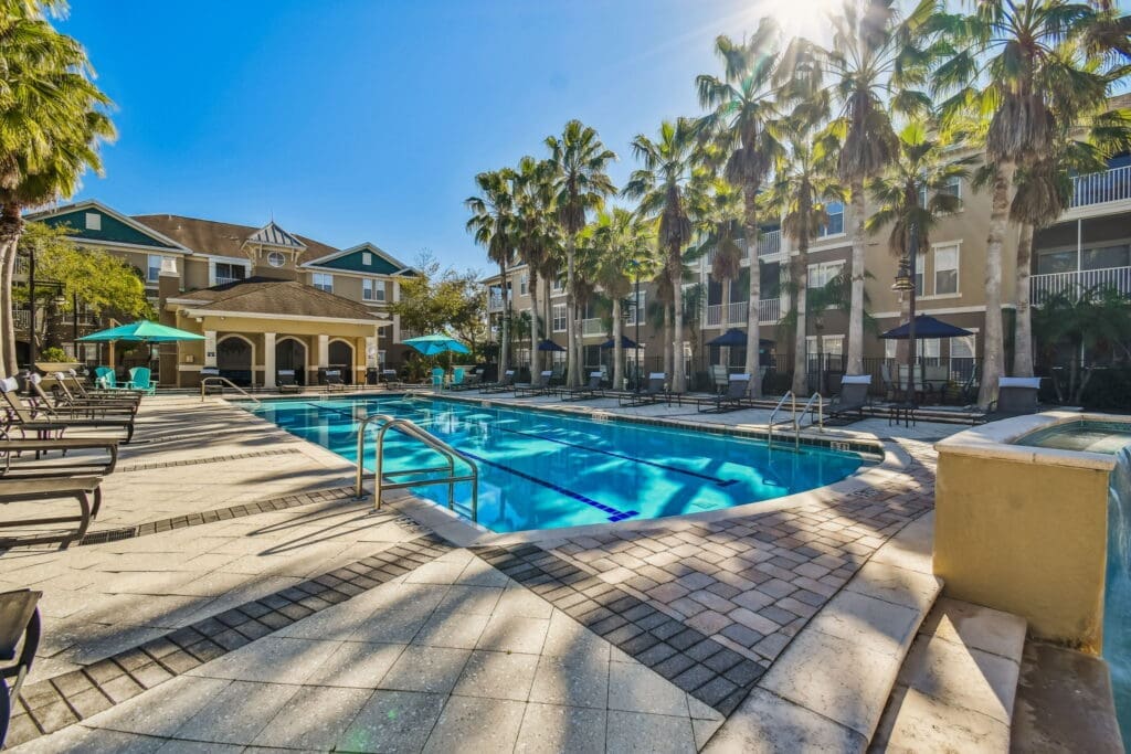 **Alt Text:** Sunny outdoor swimming pool area at an upscale apartment complex, surrounded by palm trees and tan-colored residential buildings with white balconies. The pool features lap lanes, metal handrails, and is flanked by tan paver decking with black accent lines. Lounge chairs and blue umbrellas provide seating along the sides, and a clubhouse with a covered patio is seen in the background.