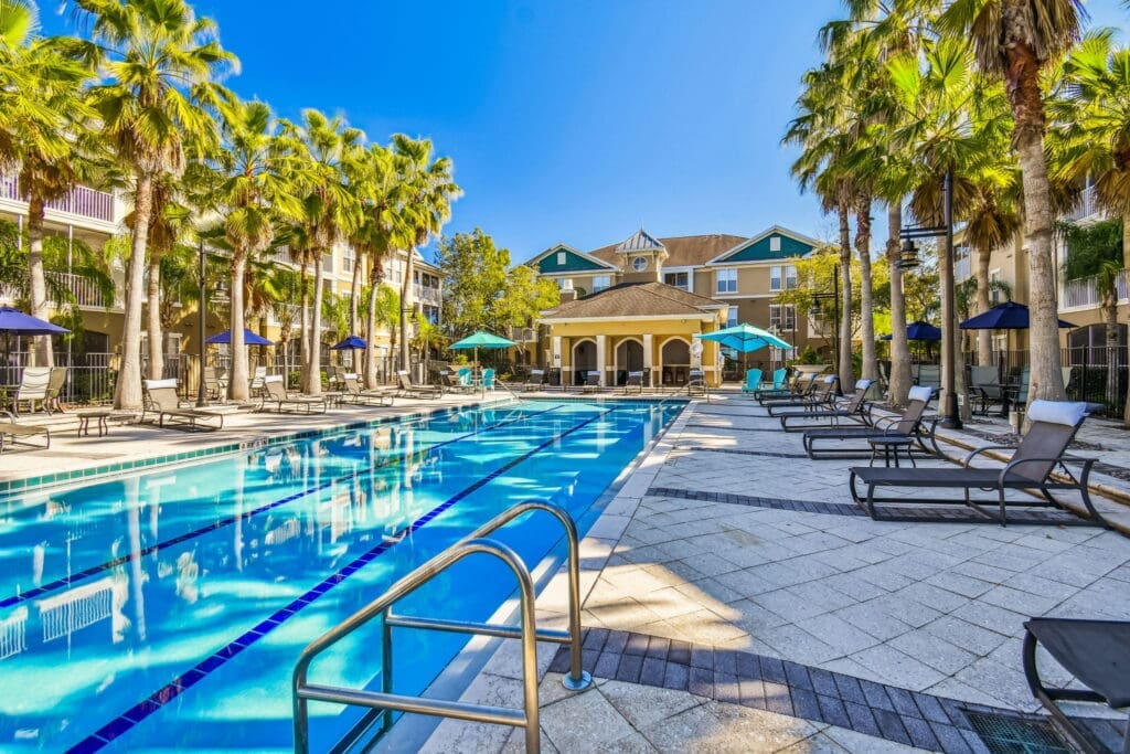 **Alt Text:** A large, resort-style outdoor swimming pool is flanked by tall palm trees and modern apartment buildings. The deck features numerous cushioned lounge chairs, umbrellas in blue and teal, and a shaded cabana area at the far end. The bright blue water and clear sky contribute to a luxurious and tropical ambiance.