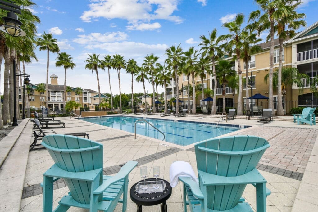 Resort-style swimming pool surrounded by tall palm trees and tan apartment buildings with balconies, featuring turquoise Adirondack chairs and black pool loungers.
