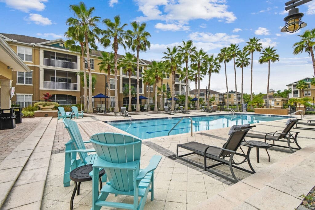 Angled view of poolside seating with Adirondack chairs and loungers along the deck of a palm-lined residential pool, surrounded by multi-level apartment buildings.