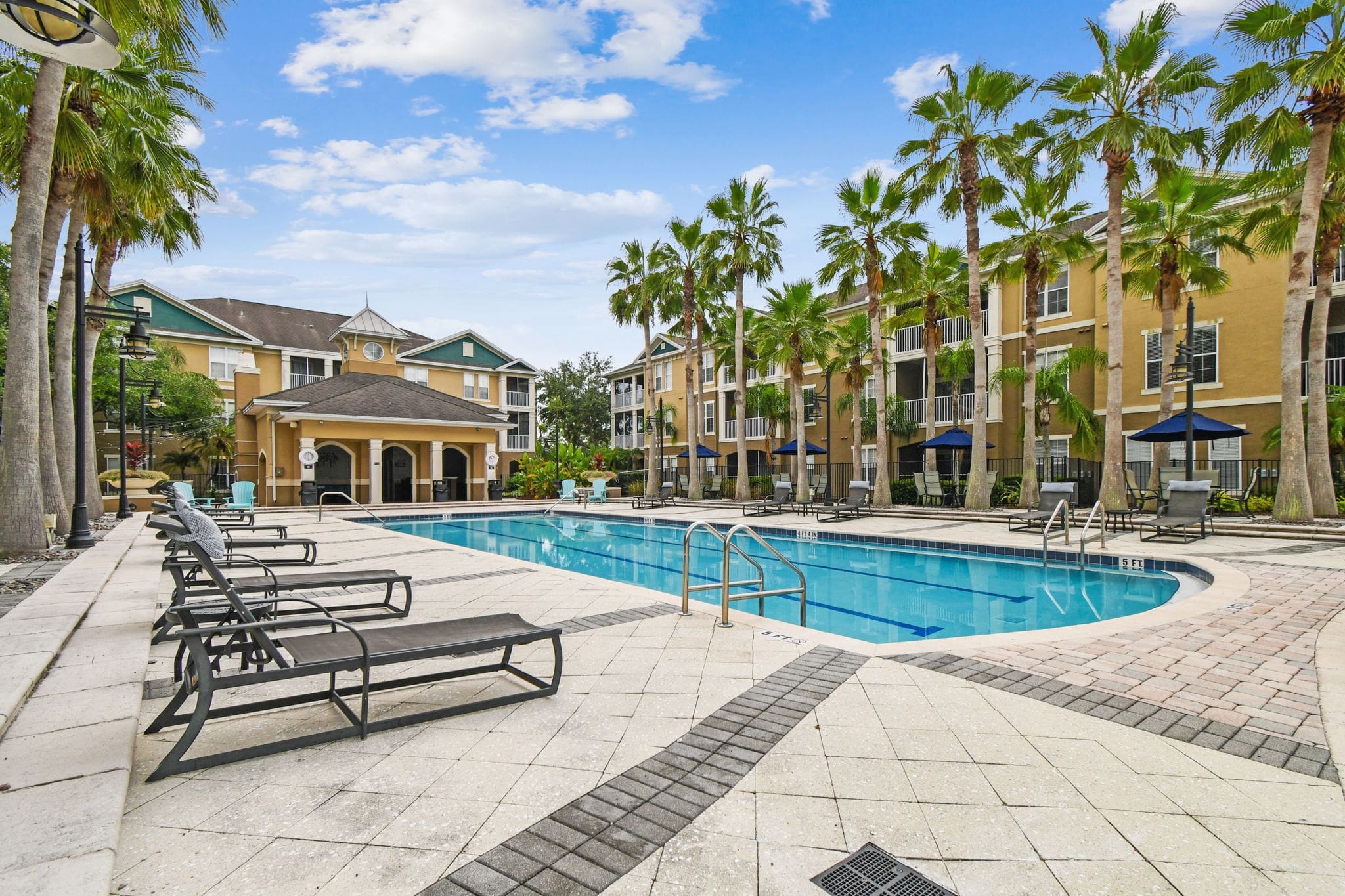 View of the pool area from one corner, highlighting the clubhouse with clock tower, apartment buildings, palm trees, and rows of pool loungers on the tiled deck.