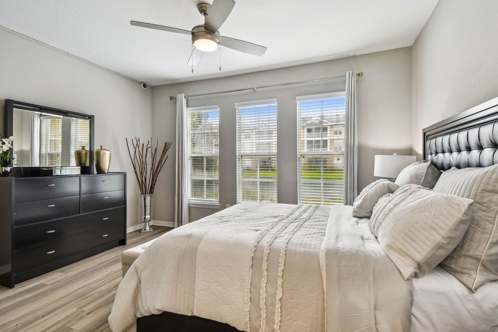 Shot from the foot of the bed showing a tall black dresser with a mirror and gold vases. A modern ceiling fan hangs above, and a silver decorative floor vase with twigs accents the corner.