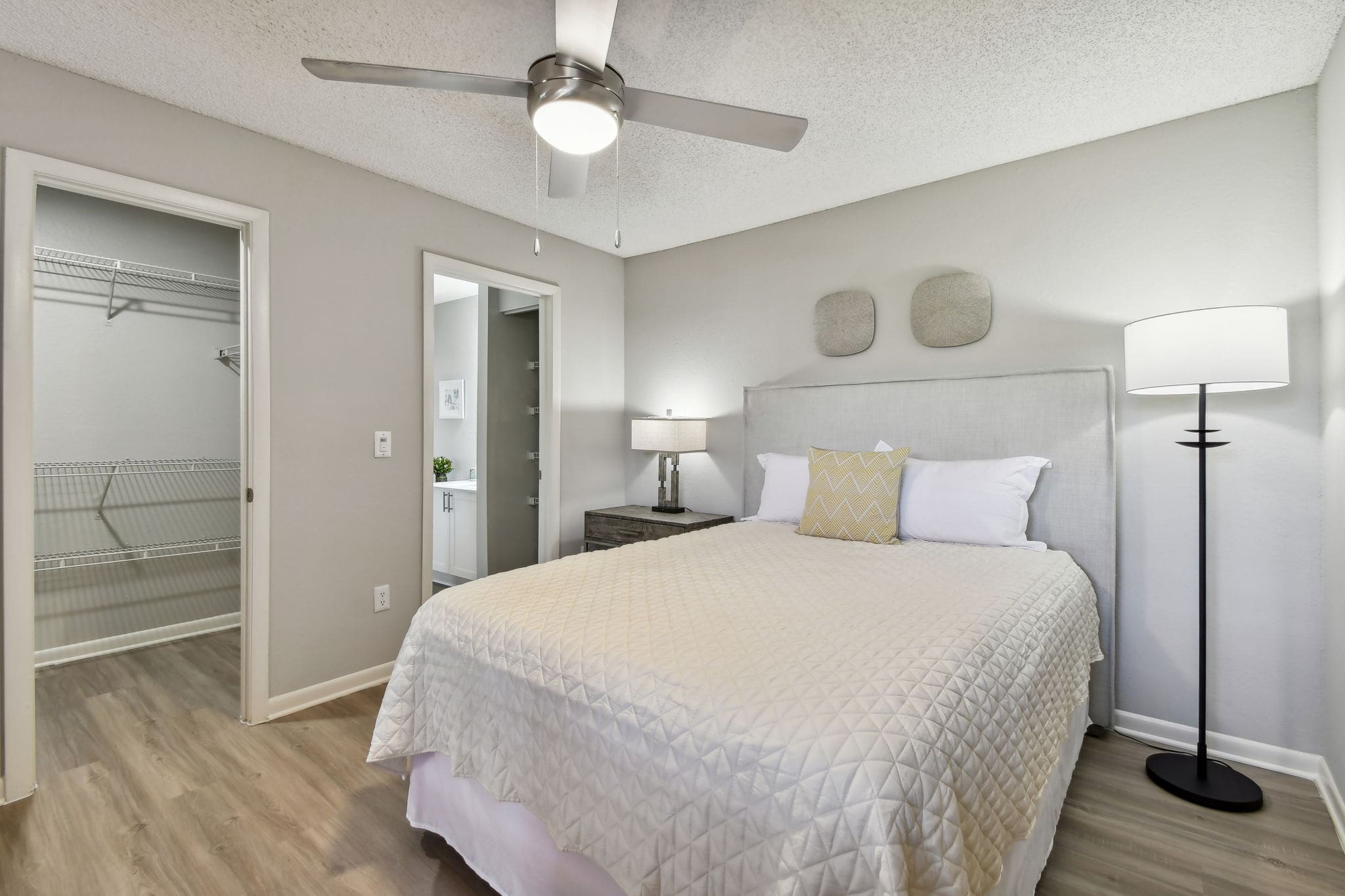 1-Bedroom Apartments in Union Park, FL - Pine Harbour - Bedroom with Wood-Inspired Flooring, Large Bed, Nightstand with Lamp, Floor Lamp, Grey Walls, Ceiling Fan, Attached Bathroom, and Spacious Closet.