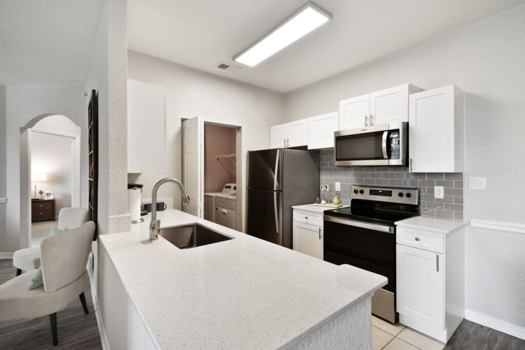 3-Bedroom Apartments In Brandon, FL - 2211 Grand Isle - Kitchen With Energy Efficient Appliances, White Cabinets, Speckled White Quartz Countertops, And In-Home Laundry Closet With Washer And Dryer.
